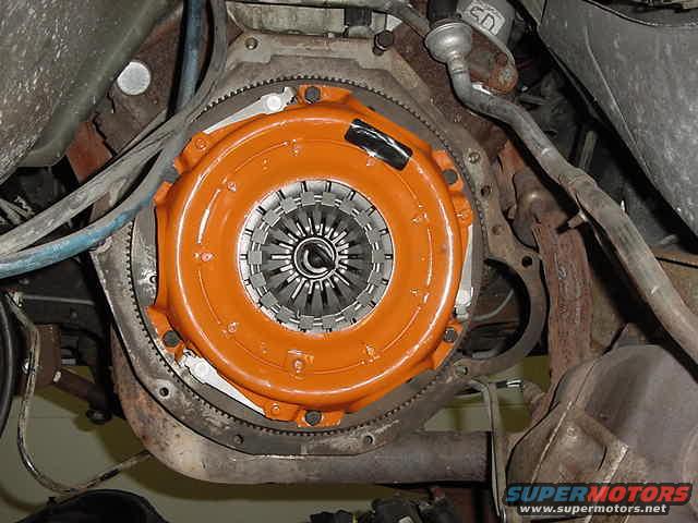 Orange clutch housing and flywheel assembly bolted to engine block, with surrounding metal components