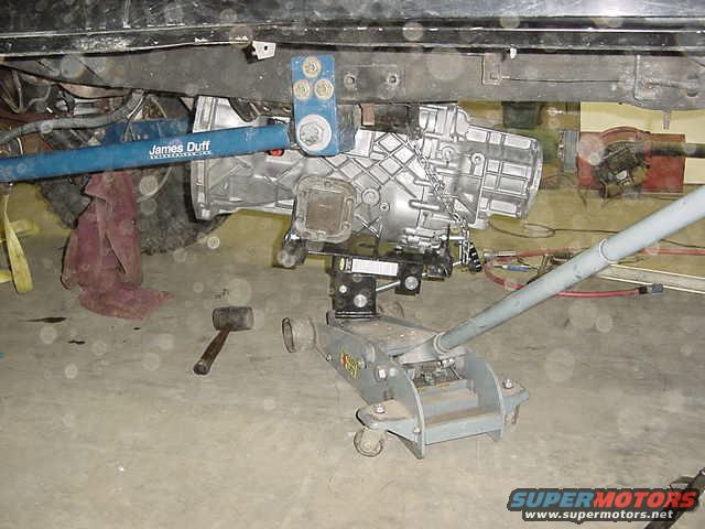 Transmission jack supporting a silver transmission with a blue James Duff suspension bar above