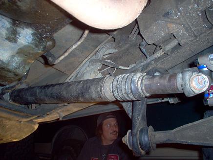 Underside of vehicle showing driveshaft, U-joint, and suspension parts. Man visible in lower right