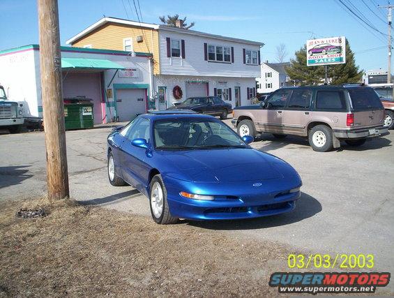 Blue 1997 Ford Probe parked outside a body shop with a tan SUV behind it