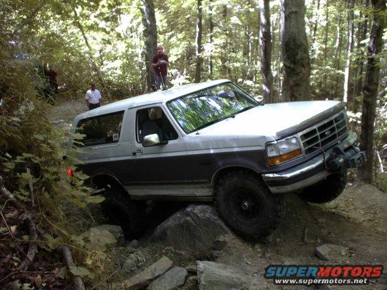 Ford Bronco navigating rocks on a wooded trail. A person stands on the roof