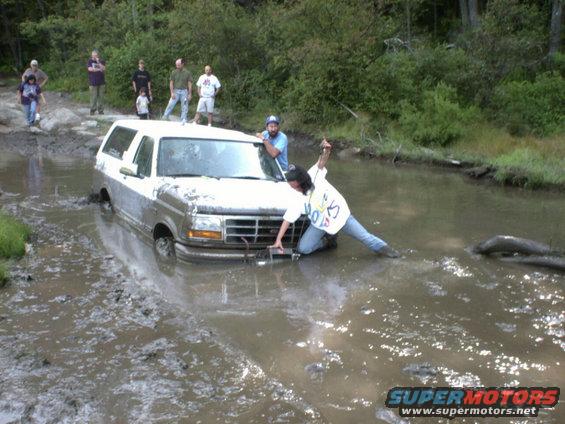 White 1995 Ford Bronco stuck in a muddy water hole with people around