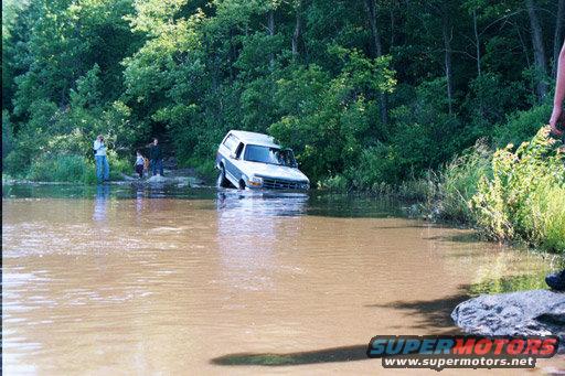 SUV partially submerged in muddy water with people watching from the bank