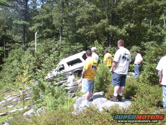 White SUV is stuck on a rocky incline in a wooded area with several people standing around it