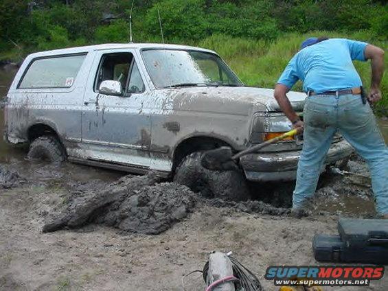 White Ford Bronco stuck in thick mud, driver digging with a shovel
