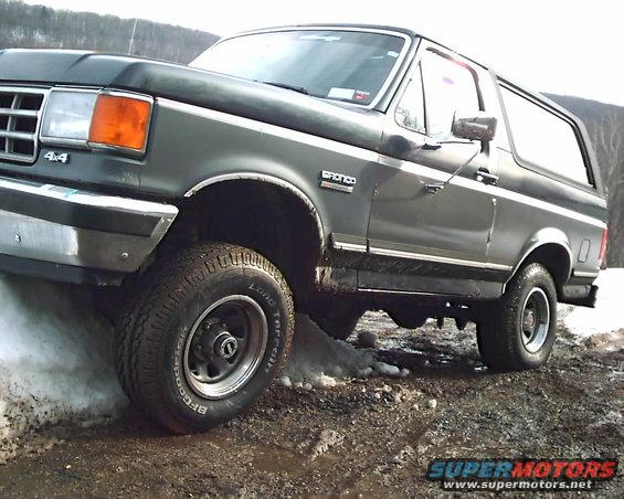 Dark green Ford Bronco on a muddy, snowy trail. The 'Bronco' badge is visible on the side