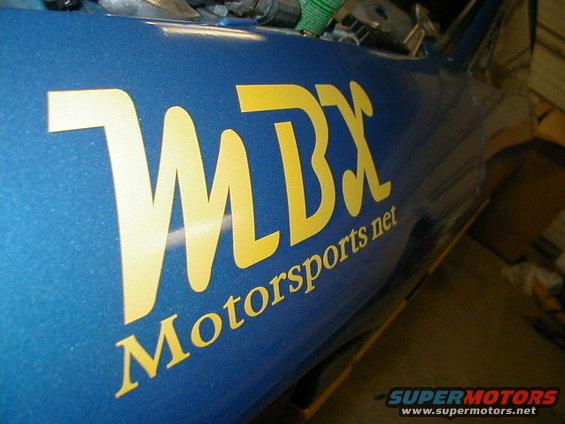 Blue car side panel with yellow MBX Motorsports net lettering. Supermotors.net logo in corner