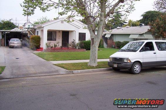 White van parked on street, sedan in driveway. House in background