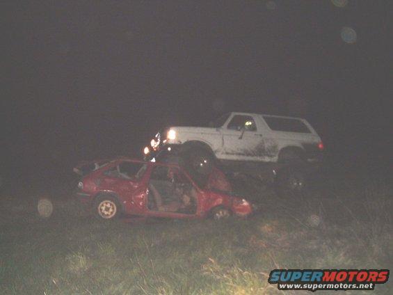 1989 Ford Bronco on top of a wrecked red car at night