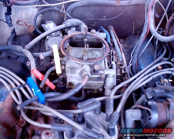 Close-up of a vehicle's engine bay, focusing on the carburetor and spark plug wires. Red and blue clothespins are attached to a hose