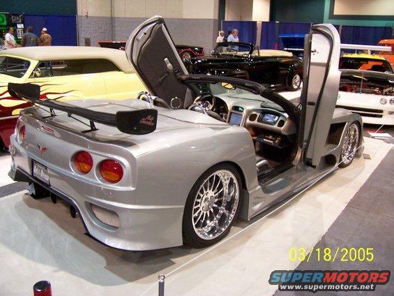 Custom silver convertible with butterfly doors, large rear spoiler, and chrome wheels. Other cars visible in background