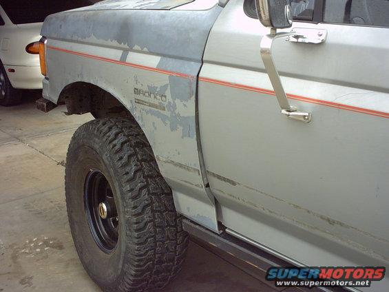 1990 Ford Bronco side view showing fender, door, and tire. Another car is visible in the background