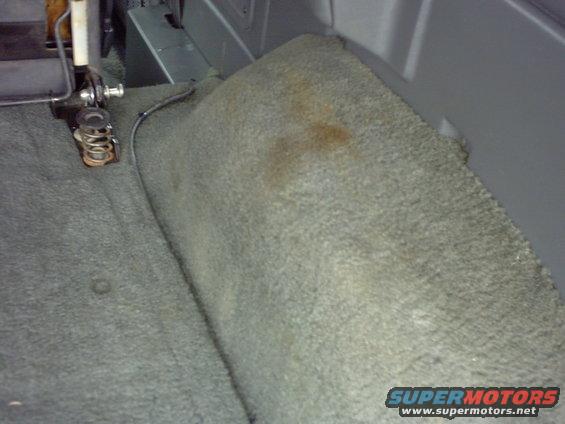 Carpeted rear interior of a vehicle, showing a seat bracket and some stains on the carpet