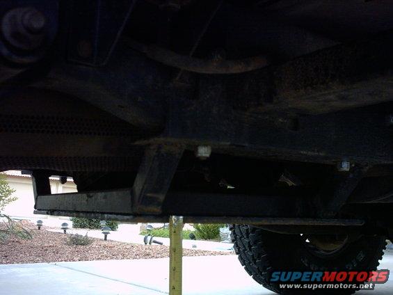 Underside view of a vehicle's frame and suspension components, with a measuring tape for scale