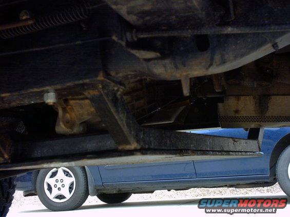 Undercarriage of a dark-colored vehicle with a blue car visible below, showing suspension components and a wheel with a star-shaped hubcap