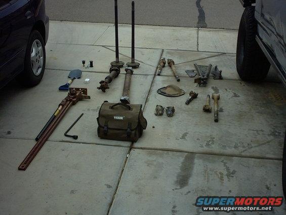 Various vehicle parts including axles, driveshafts, and a Hi-Lift jack on a driveway