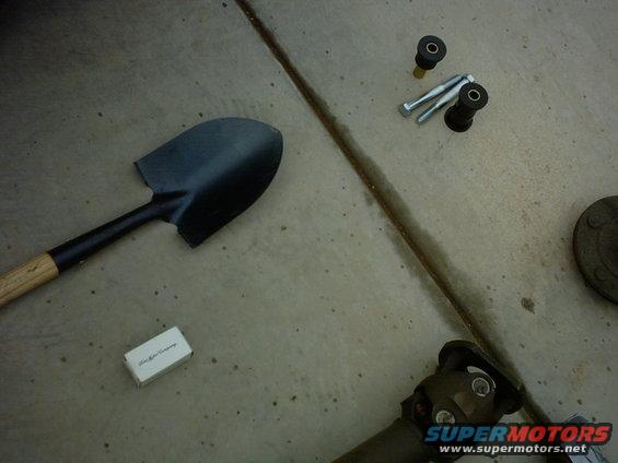 Shovel, bolts, bushings, and a driveshaft U-joint on concrete. Small white box has text