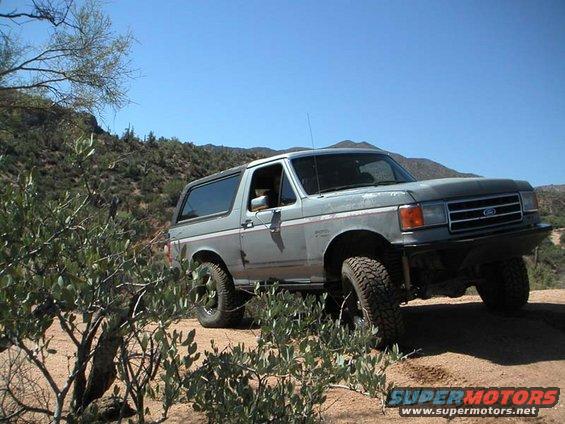 1990 Ford Bronco on a dirt trail with desert vegetation and mountains in background