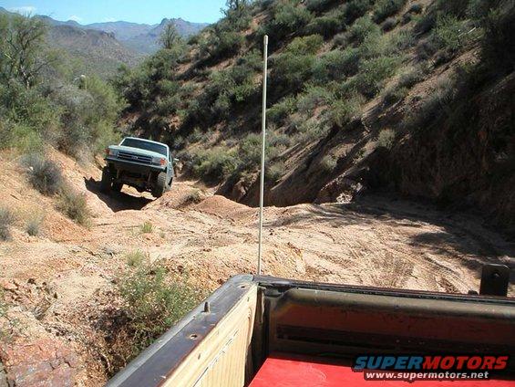 Light-colored Ford Bronco climbing a dirt hill, viewed from the bed of another vehicle