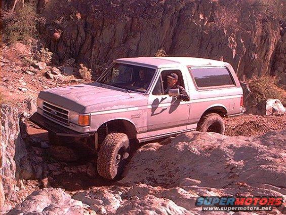 1987 Ford Bronco navigating a steep, rocky trail with one front wheel lifted