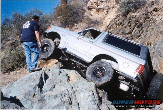 1990 Ford Bronco climbing over large rocks on a trail, driver standing nearby