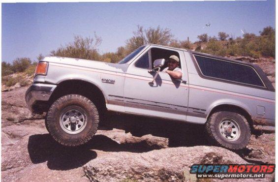 Silver 1990 Ford Bronco driving over rocks on a sunny day