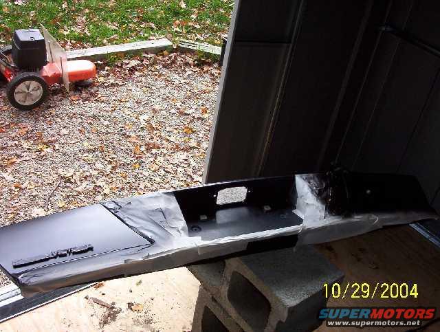 Black car bumper on cinder blocks, partially covered in paper. Lawn mower in background