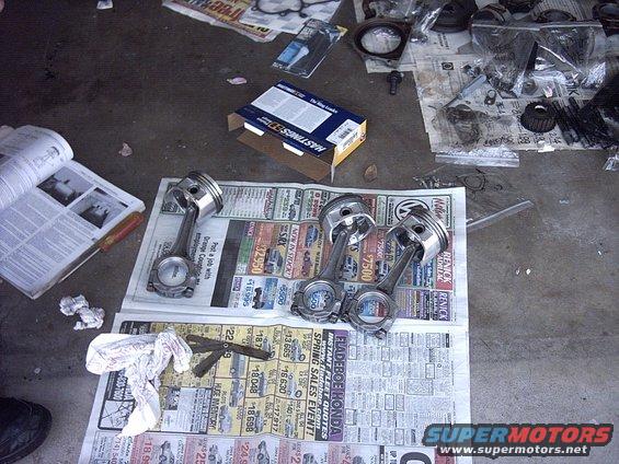 Engine pistons and connecting rods laid out on newspapers, with a manual and parts box nearby