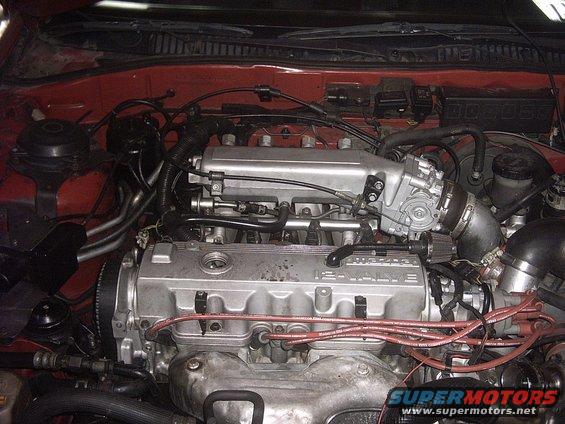 Engine bay with a red car exterior visible, showing a 16-valve engine with silver intake manifold and red spark plug wires