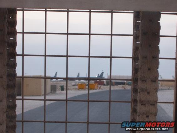 Airplanes on tarmac viewed through a metal grate, with buildings and bins in the background