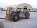 1988 Ford Bronco - A tan forklift with heavy-duty tires sits in a lot near a large arched building, with other vehicles visible in the background.