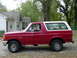1988 Ford Bronco - This red Ford Bronco with a white top is seen from the side, parked on a gravel surface next to a house.