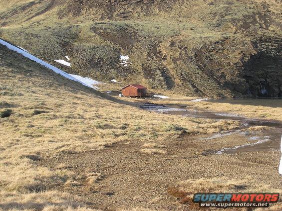 Small wooden cabin with a red roof sits in a valley with dry grass, snow patches, and a stream