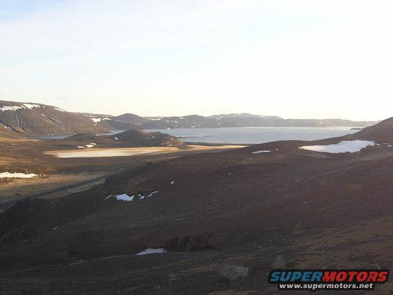 Vast Icelandic landscape with mountains, a lake, and snow patches under a bright sky