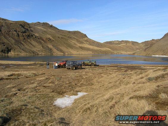 Three off-road vehicles parked near a lake with mountains in the background