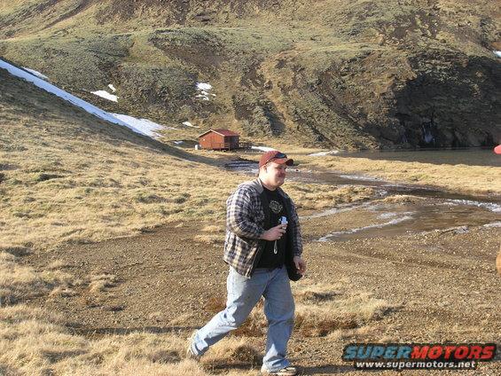 Man in plaid shirt and jeans walks through dry grass with a small cabin and hills in the background