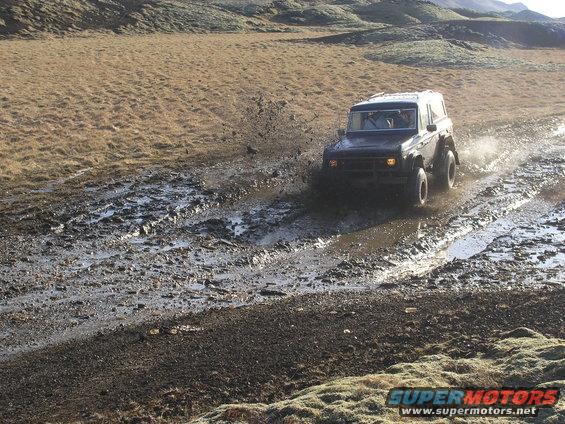 Dark SUV driving through mud, splashing water. Visible text 'SUPER MOTORS'