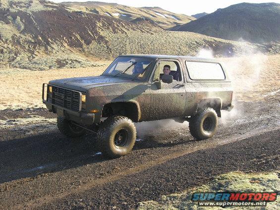 Camouflaged off-road SUV with lifted suspension driving on a dirt track, kicking up dust. Driver visible