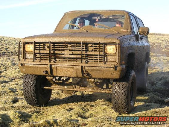 Mud-covered SUV with a large grille and off-road tires driving on a grassy, uneven terrain