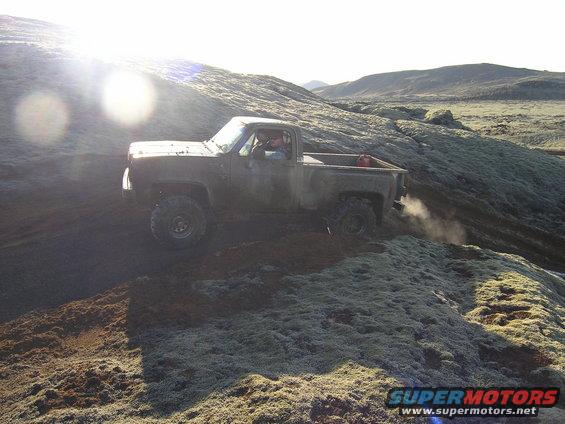 Pickup truck driving off-road on a mossy, rocky incline. A red gas can is visible in the truck bed