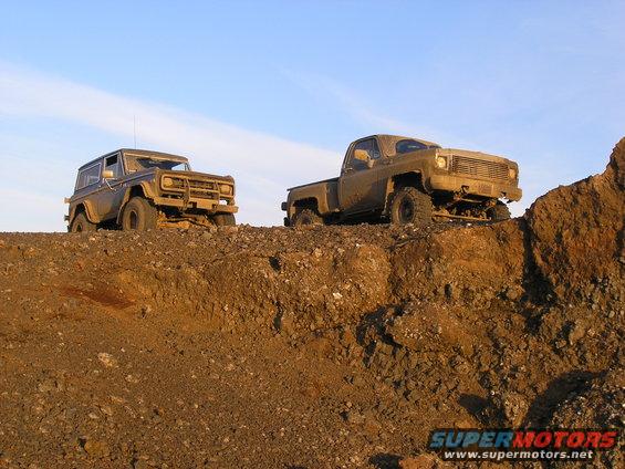 Two muddy off-road vehicles, a Bronco and a pickup truck, are positioned on a dirt mound under a clear sky