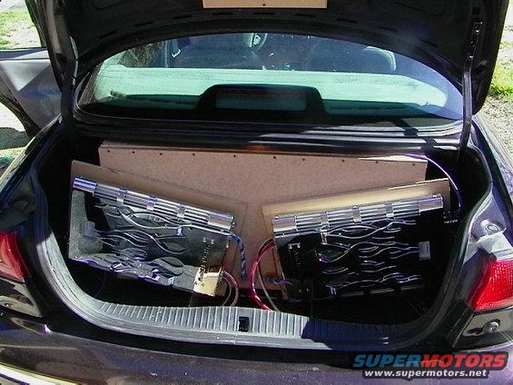 Car trunk with two amplifiers mounted on a wooden board, wires visible