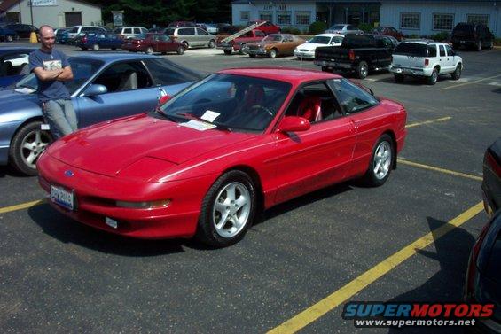 Red 1993 Ford Probe coupe parked in a lot, front quarter view, with a man standing next to a blue car
