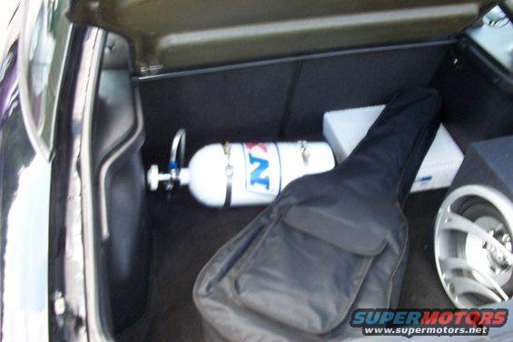 Car trunk holding a white nitrous bottle with blue lettering, a black guitar case, and a white box