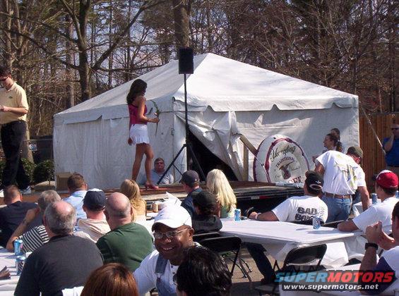 People gathered outdoors around tables, with a tent and stage set up. A woman is on the stage