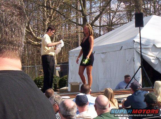 People on a stage with a tent and audience, a man in a yellow shirt reads from paper, a woman in a black dress holds flowers