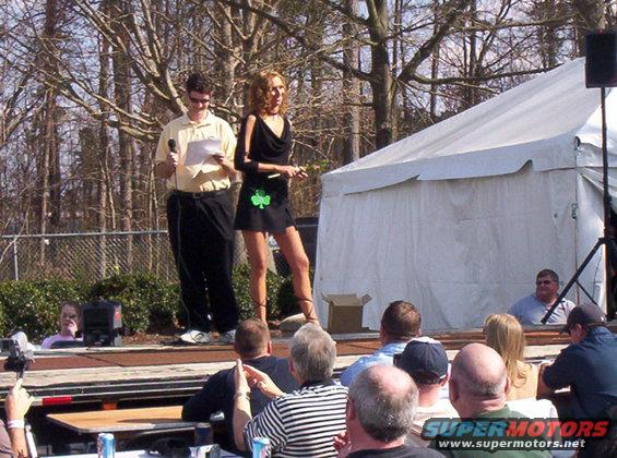 Man holding papers and woman in black dress on outdoor stage with audience and tent