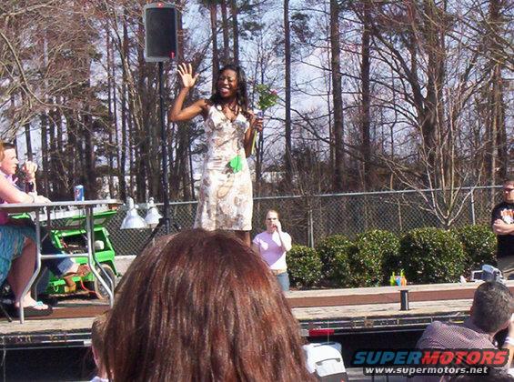 Woman in a patterned dress waves on a stage with trees and a fence in the background