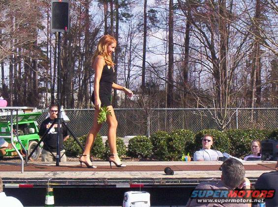 Woman in black dress walks on stage holding greenery. A green utility vehicle is visible to the left