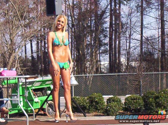 Woman in teal bikini and green beaded necklace on stage with a green lift in the background